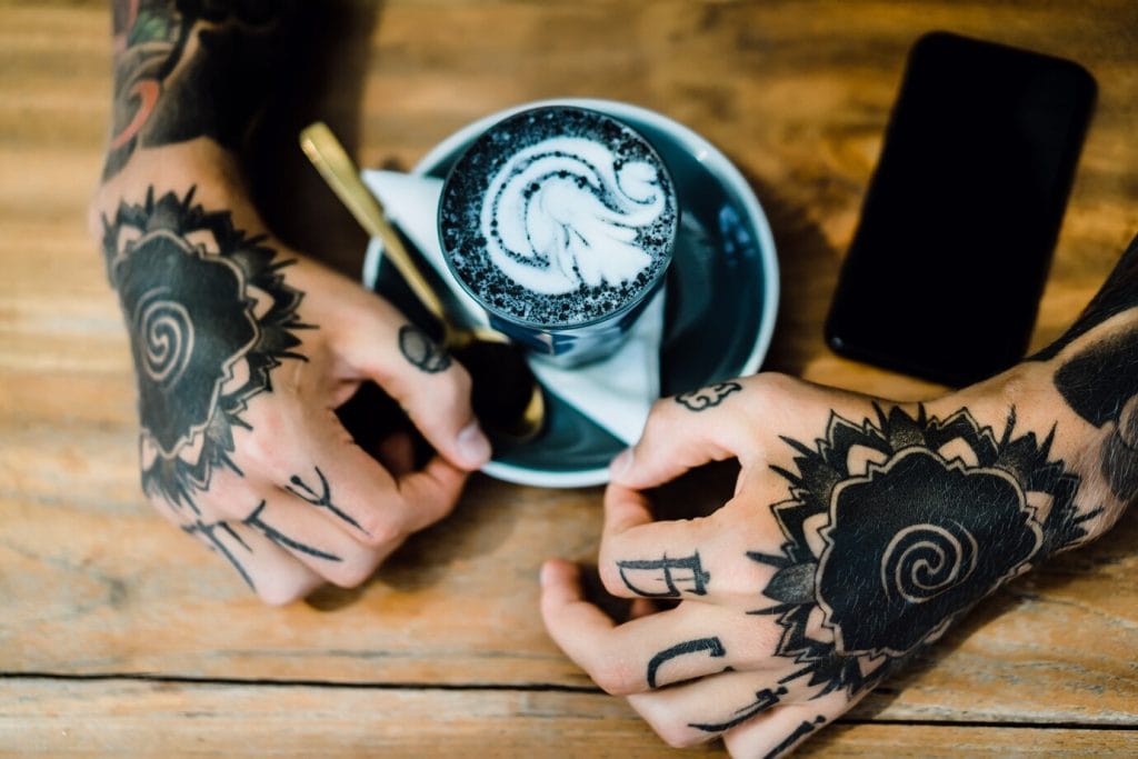 Cafe cu latte art, tatuaje pe mâini și telefon pe masă la Old Ink Tattoo București, studio de tatuaje profesionist, design unic și servicii de top în tatuare în București.